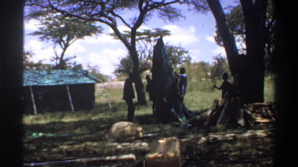 1969 Lagos, Nigeria home-movie frame with several people around a raised tarp, poles, trees, grass, and a small blue-roofed structure.
