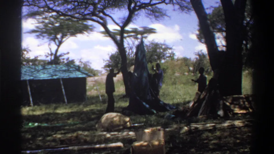 In Lagos, Nigeria, 1969 home-movie footage, several men appear near a blue tarp or tent under trees beside a low roofed structure.