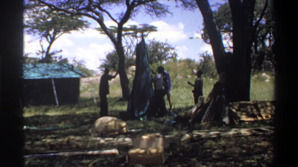 In 1969 Lagos home-movie footage, several men appear to work around a dark tent or awning beneath shade trees in a grassy setting.