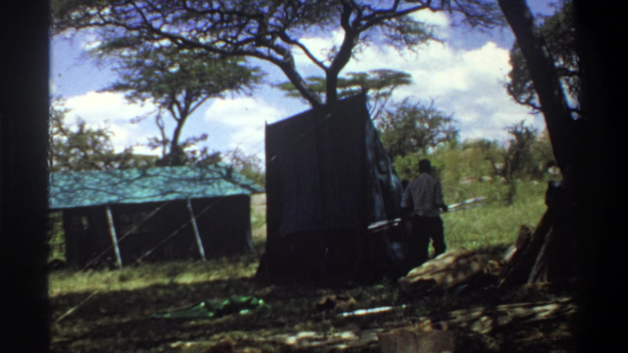 1969 Lagos 8mm frame showing adults near a dark upright shed panel, a low green corrugated roof, grass, and shade trees.