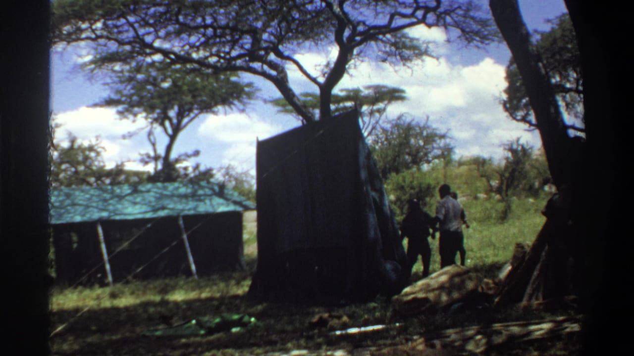 1969 Lagos 8mm frame showing two adults beside a dark shed panel, with a low roofed structure, grass, and trees visible.