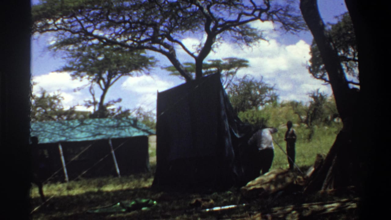 1969 Lagos, Nigeria frame showing two people near a dark tent panel, with trees and a green-roofed shelter in the background.