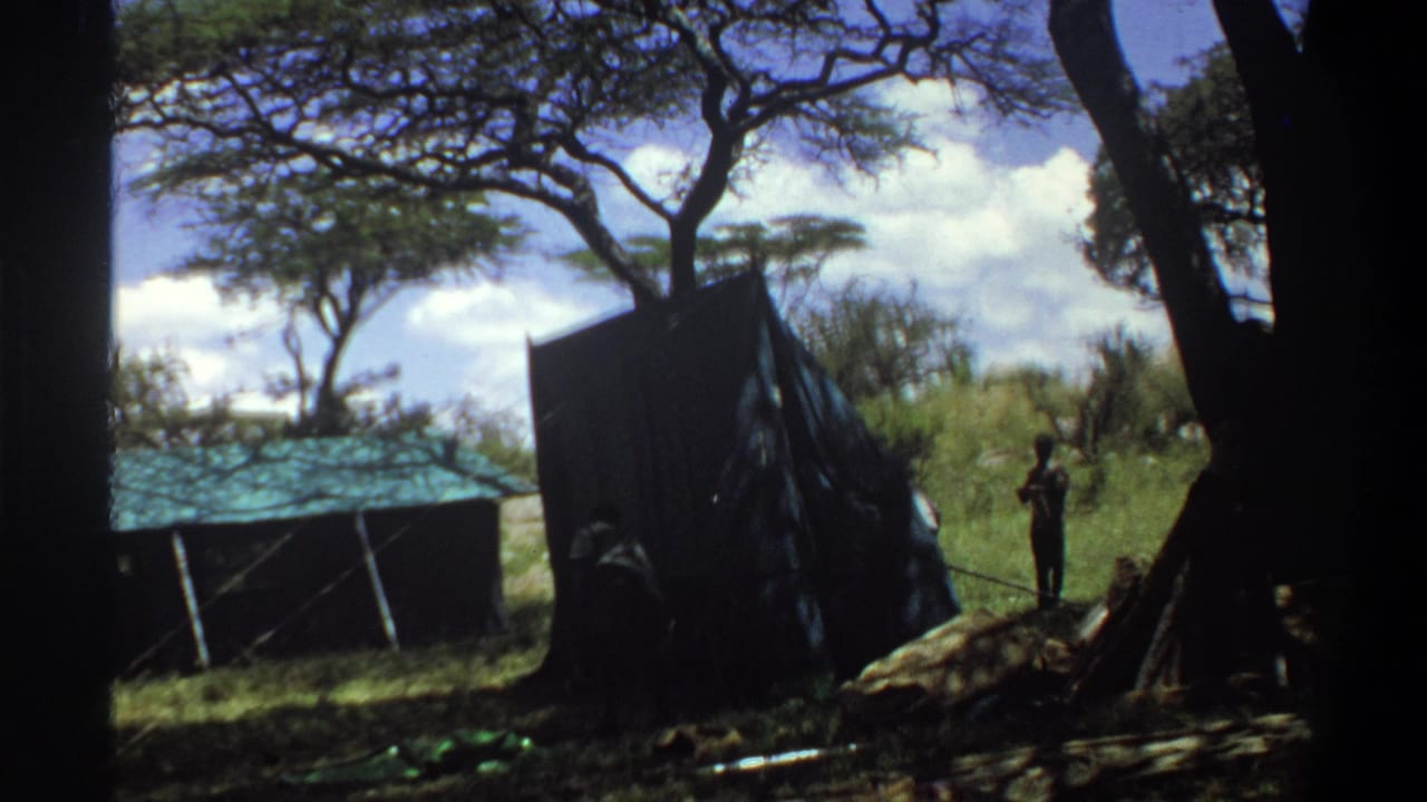1969 Lagos-area 8mm frame showing people near a dark truck, field shelter, trees, and supplies on grassy ground.