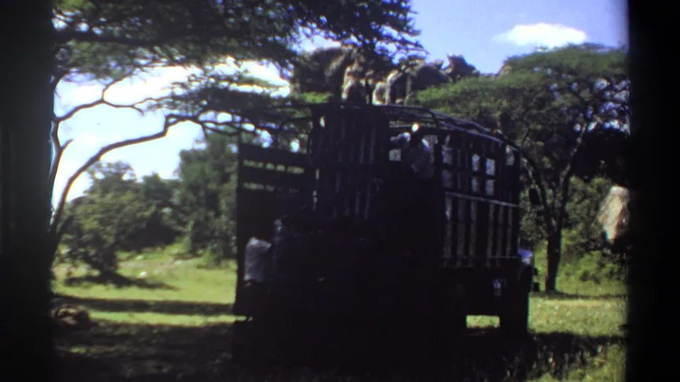 1969 Lagos home-movie frame showing a large open-bed truck under trees, with people visible in the rear against bright grass and sky.