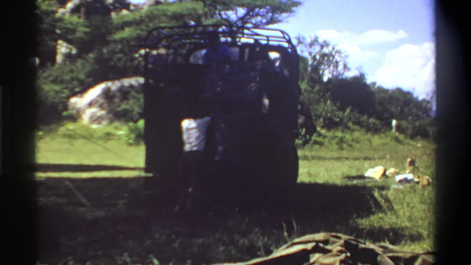 1969 Lagos, Nigeria home-movie frame with a shaded rear view of a truck, passengers faintly visible, and luggage-like objects in the grass.