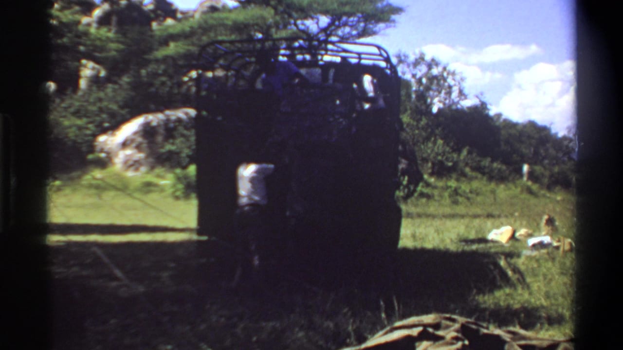 1969 Lagos home-movie frame showing a dark roof-rack truck on grass, people in shade, trees, rock, and bright sky.