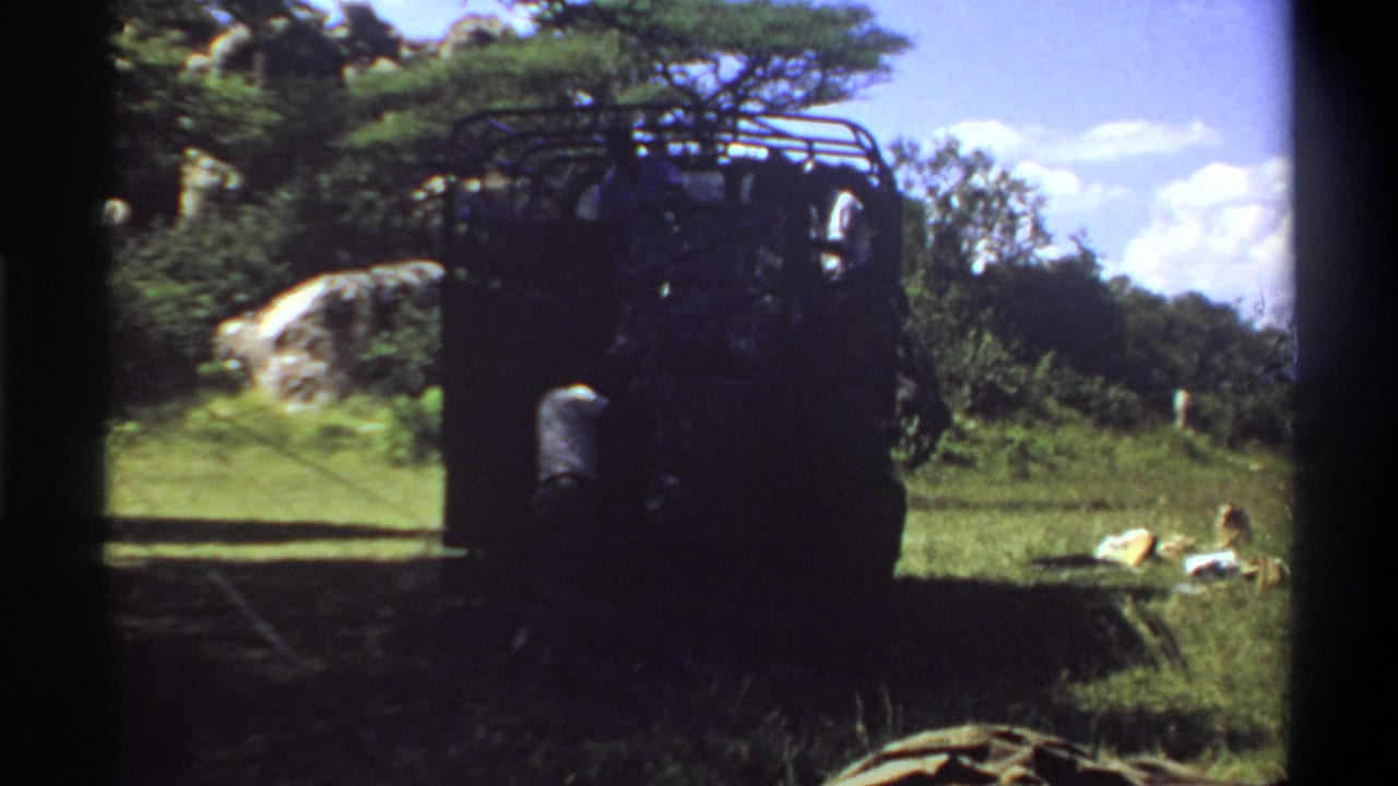 1969 Lagos footage frame with a dark truck and roof rack in partial shade, grassy foreground, trees, and items on the ground.