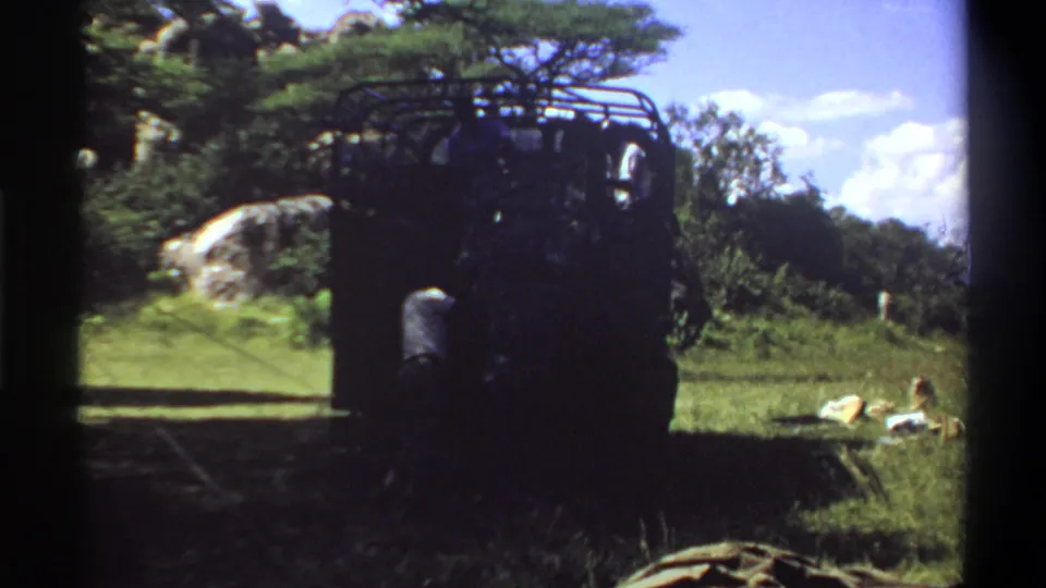 1969 Lagos footage frame with a dark truck and roof rack in partial shade, grassy foreground, trees, and items on the ground.