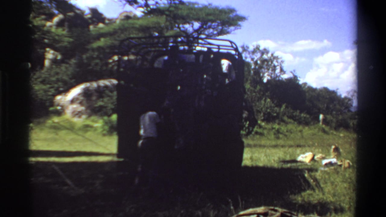 1969 Lagos frame showing a man in a light shirt near the rear of a shaded truck, with grass, trees, rock, and bright sky visible.