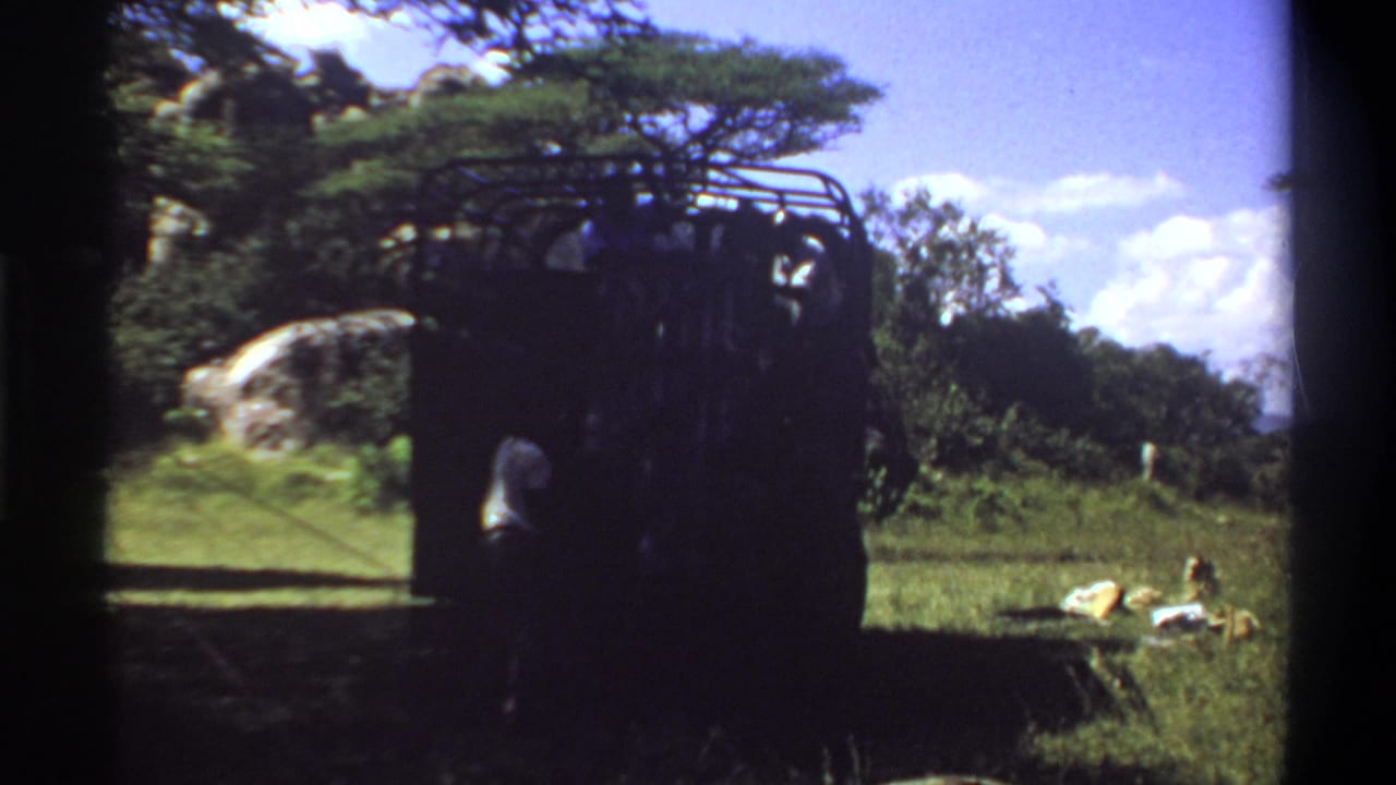 1969 Lagos, Nigeria home-movie frame of a dark safari-style truck with a person nearby, set among grass, trees, rock, and bright sky.