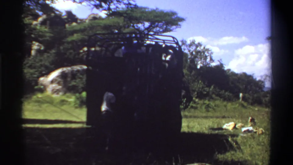 1969 Lagos, Nigeria home-movie frame of a dark safari-style truck with a person nearby, set among grass, trees, rock, and bright sky.