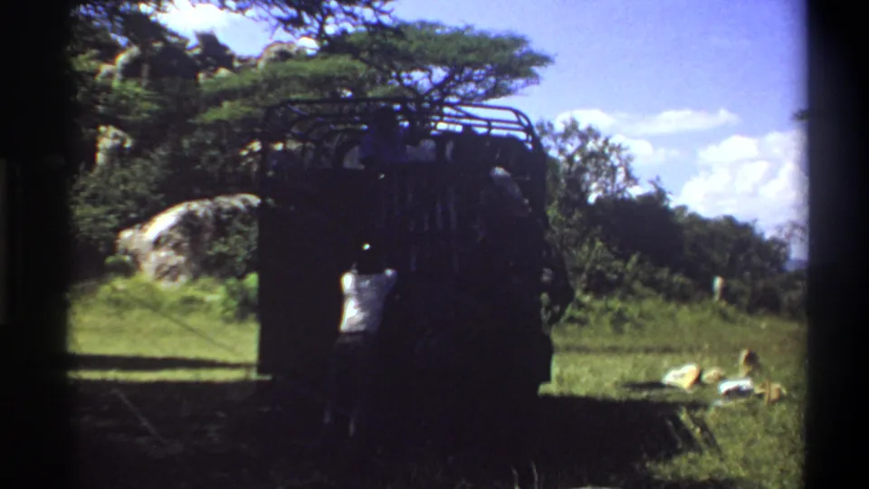 1969 Lagos home-movie frame showing a dark roof-rack truck in grass with a man in a light shirt near the rear.