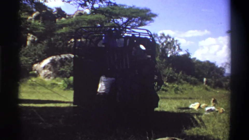A man appears behind a dark safari-style truck in 1969 Lagos footage, with trees, rocks, and bright sky beyond.