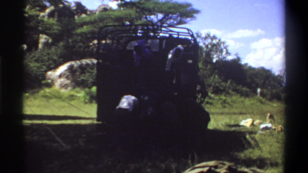 1969 Lagos, Nigeria home-movie frame showing men near a dark safari vehicle in a grassy outdoor area with rocks and trees.