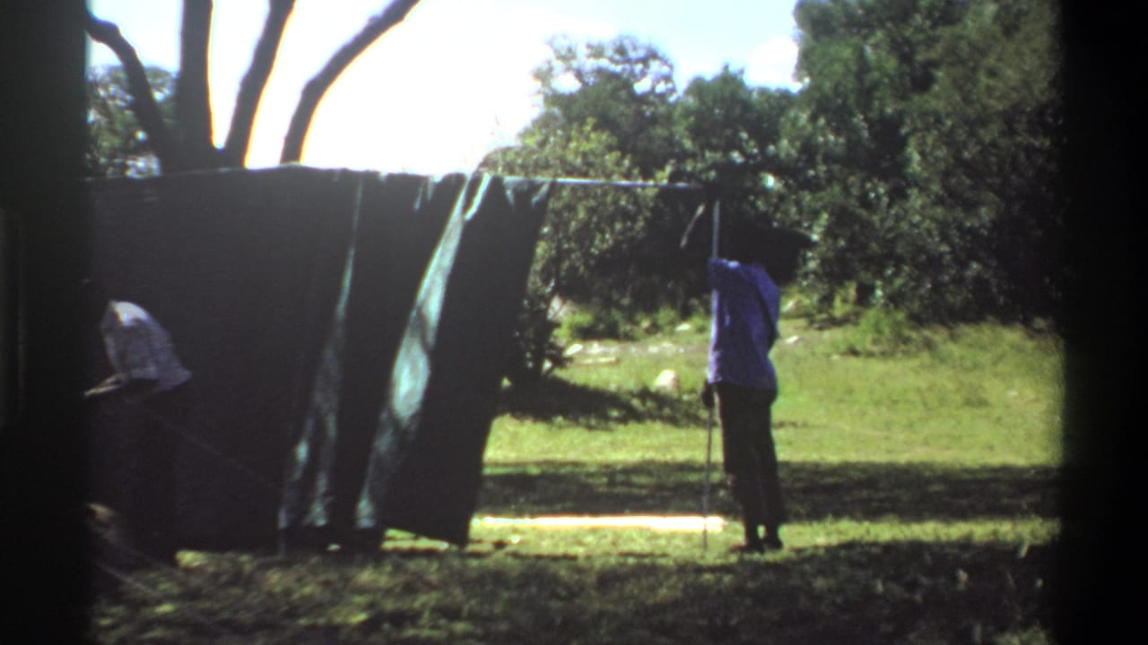 1969 Lagos, Nigeria frame showing men erecting canvas tent panels with poles and ropes in a grassy, tree-lined area.