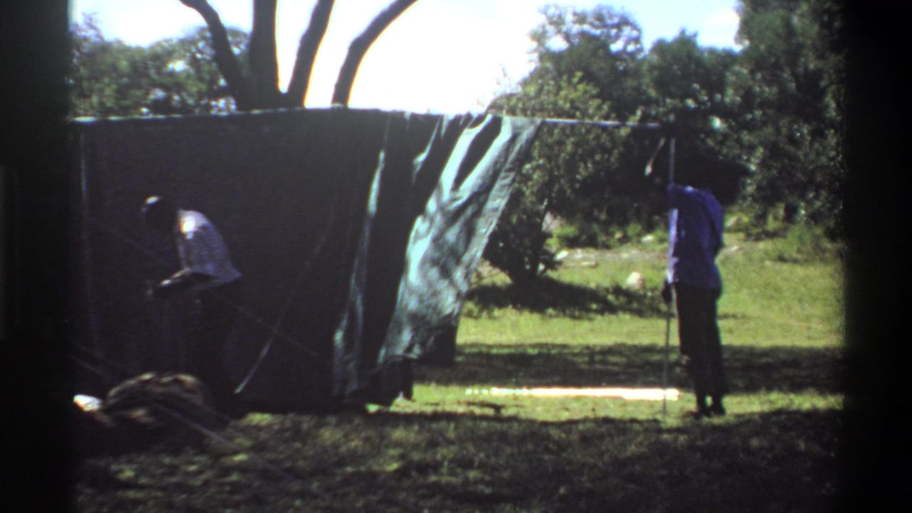 In Lagos, Nigeria home-movie footage from 1969, two people appear to adjust a large dark tarp or tent wall in a grassy outdoor area.