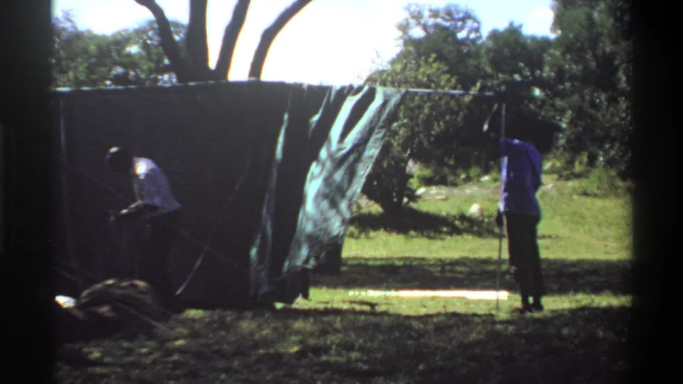 In Lagos, Nigeria home-movie footage from 1969, two people appear to adjust a large dark tarp or tent wall in a grassy outdoor area.