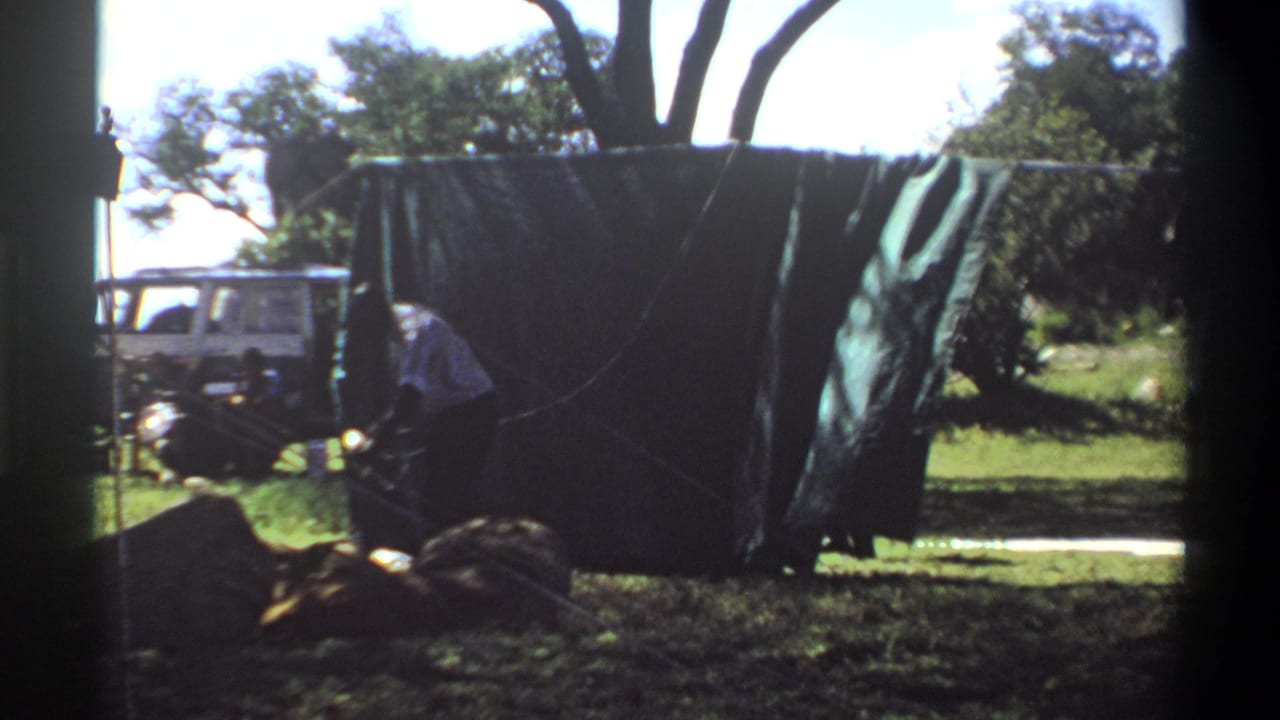 A person works low near the base of a dark tarp or tent, with a white utility vehicle visible in the background of the Lagos 1969 home movie.