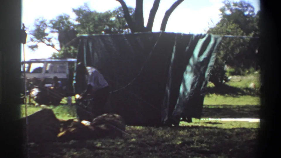 A person works low near the base of a dark tarp or tent, with a white utility vehicle visible in the background of the Lagos 1969 home movie.