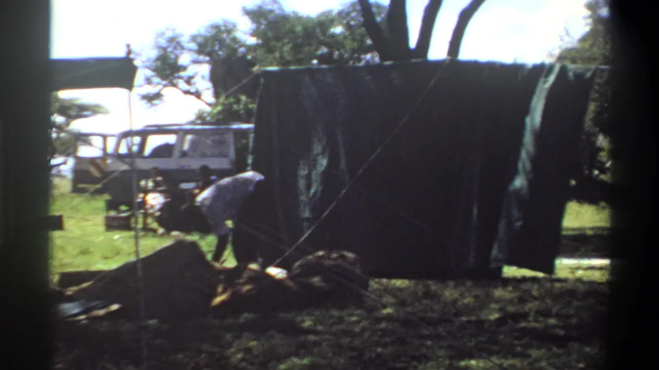 The frame shows a person adjusting material beside a large dark tarp, with a white vehicle, trees, grass, and poles visible in the 1969 Lagos footage.