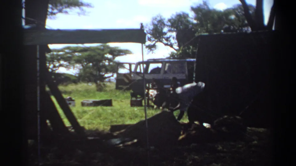 1969 Lagos home-movie frame showing a grassy campsite with trees, a white utility vehicle, dark shelter, and a man working near wood.