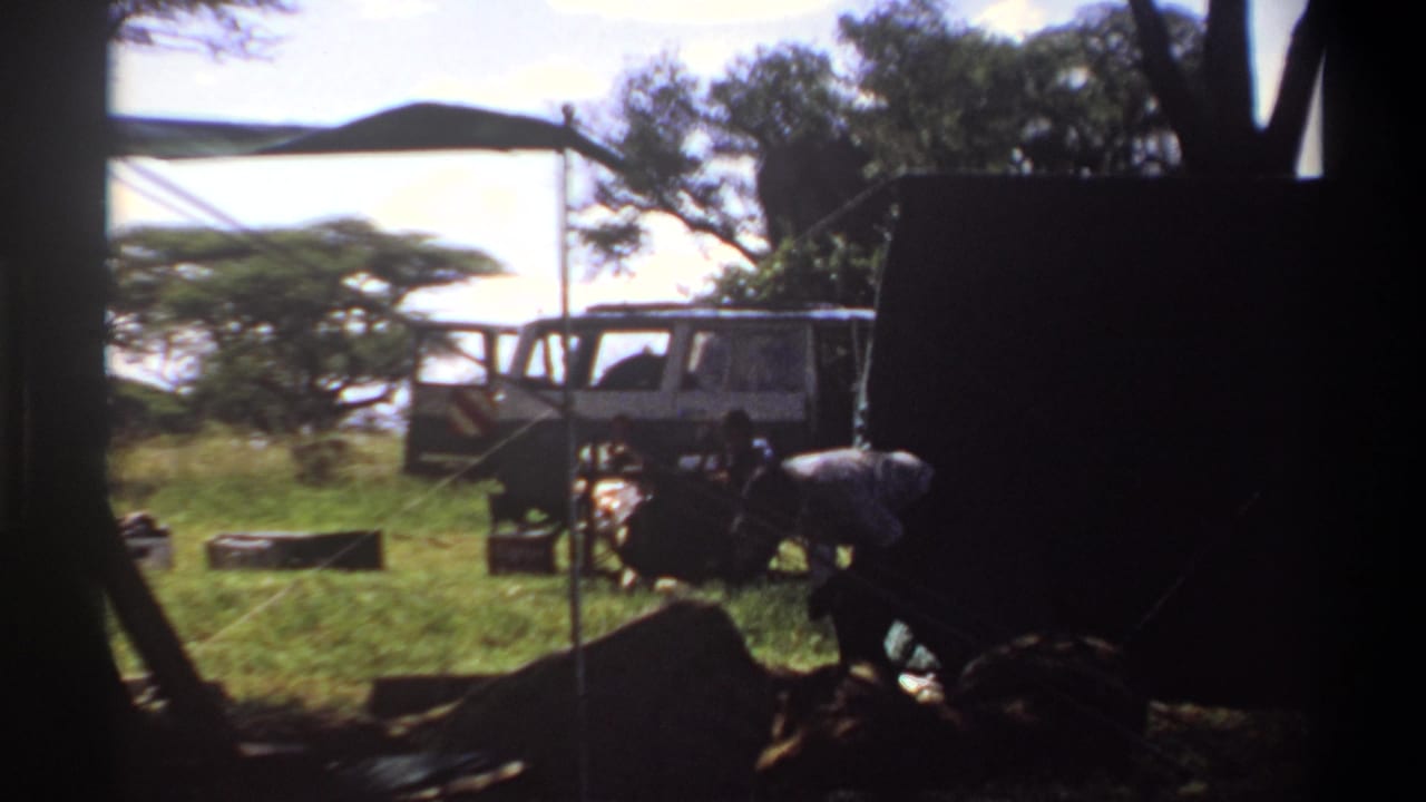 A 1969 Lagos home-movie frame shows a shaded campsite opening onto grass, trees, camp gear, a white vehicle, and a man bending at work.