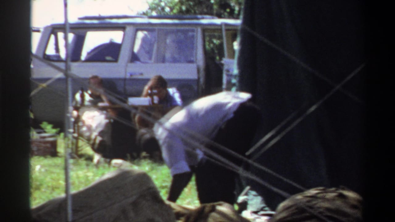 People near a dark tent structure with a white van parked behind them in a grassy field.