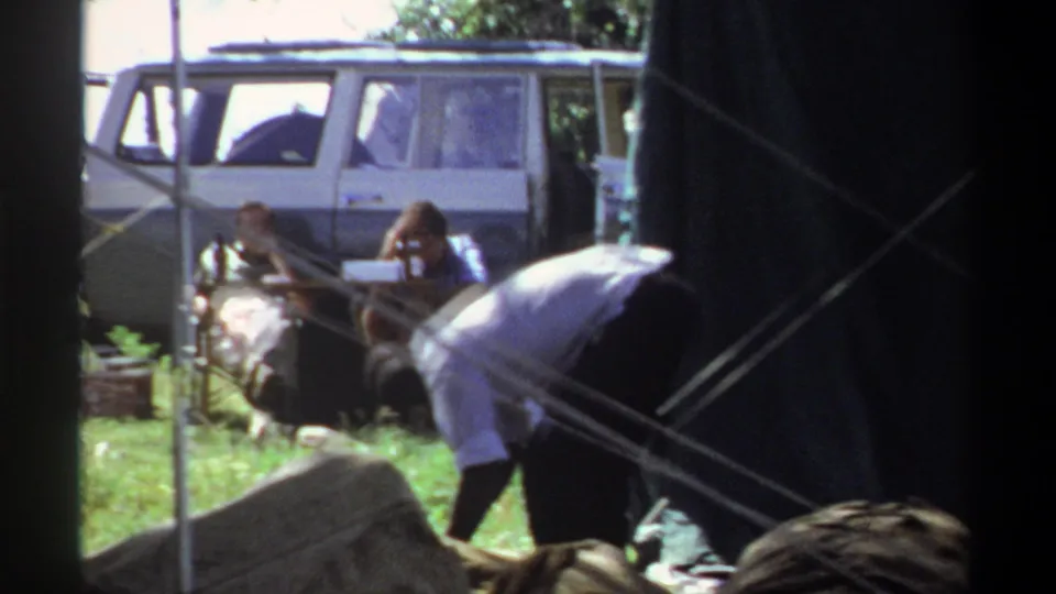 1969 Lagos home-movie frame showing people beside a large dark tent, crossing ropes, and a white van in a grassy outdoor area.