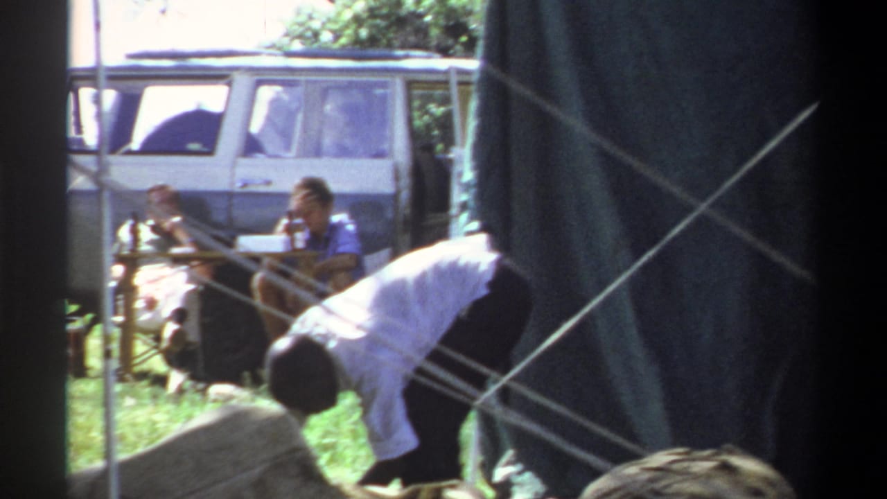 A person bends near tent ropes while others sit near a parked white van.