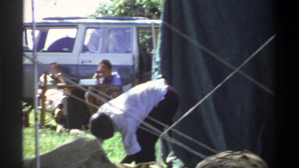 1969 Lagos frame with a person in a white shirt adjusting the tent area as seated adults and a white van appear behind crossed ropes.