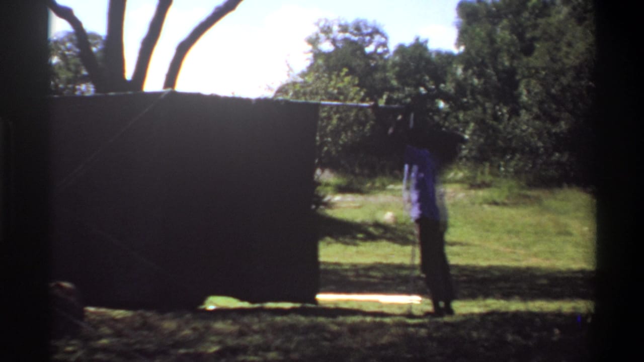 1969 Lagos frame with a man beside a dark outdoor screen or tarp, with grass, trees, and bright daylight in the background.