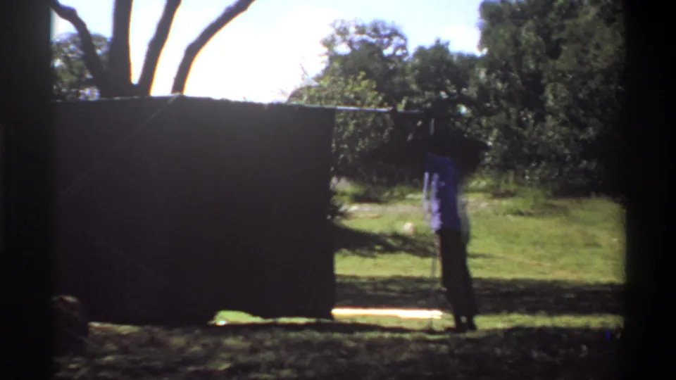 1969 Lagos frame with a man beside a dark outdoor screen or tarp, with grass, trees, and bright daylight in the background.