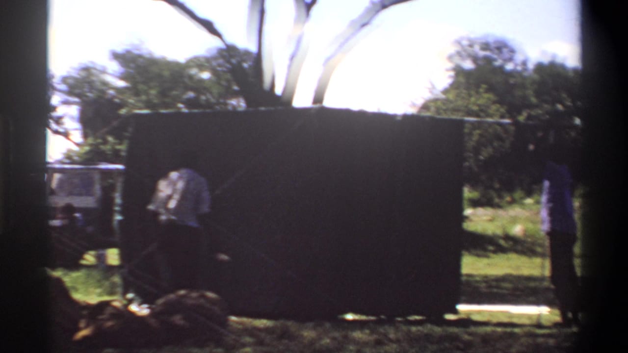 1969 Lagos home-movie frame showing two people around a large dark tarp or curtain, with a white vehicle partly seen behind trees.