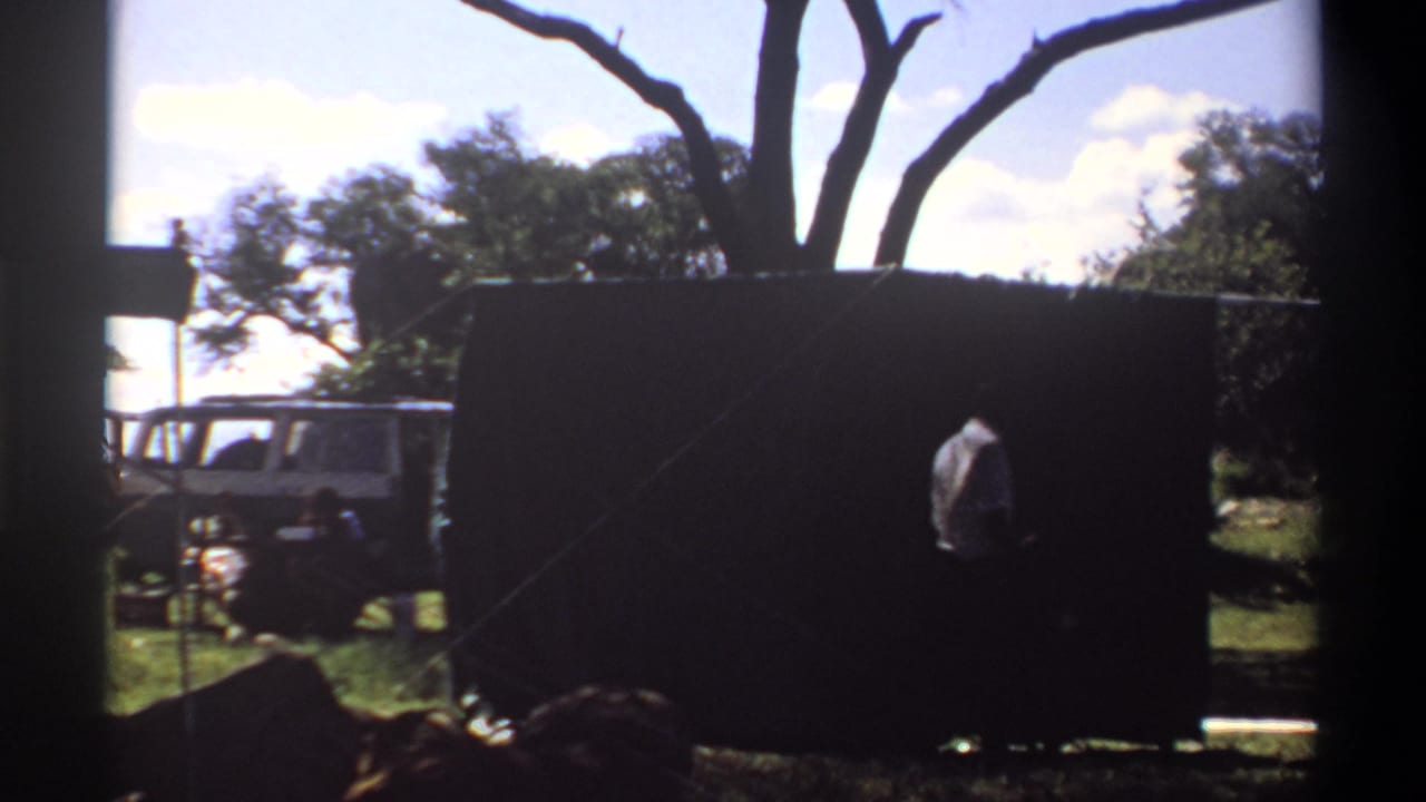 1969 Lagos frame with a person near a large dark curtain or tarp, a white vehicle behind it, and trees under bright daylight.
