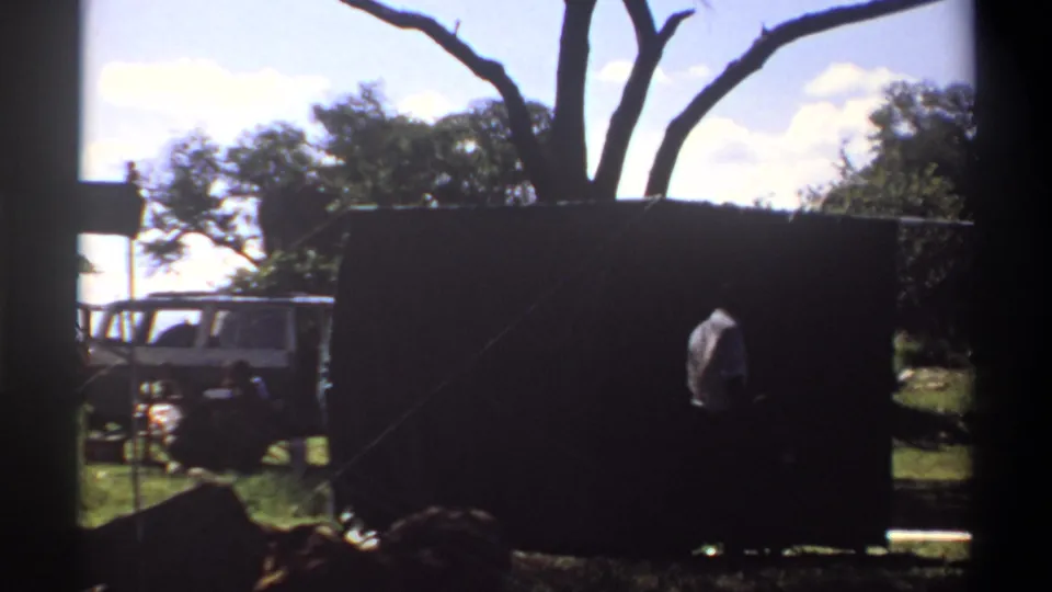 1969 Lagos frame with a person near a large dark curtain or tarp, a white vehicle behind it, and trees under bright daylight.