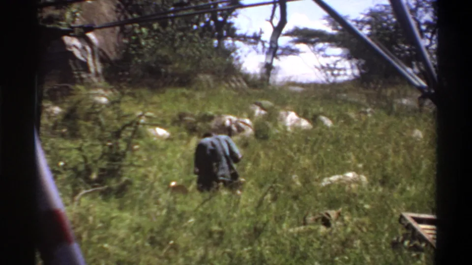 Soft 8mm frame from Lagos, 1969, with a man bent forward in dense grass and pale rocks visible across the background.