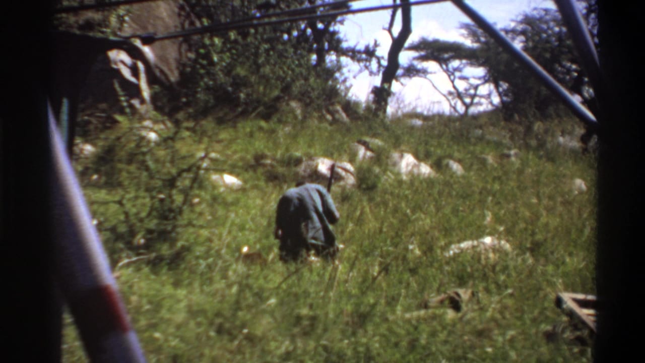 1969 Lagos home-movie still showing a rear view of a man in tall grass, framed by dark structural elements with rocks and trees beyond.