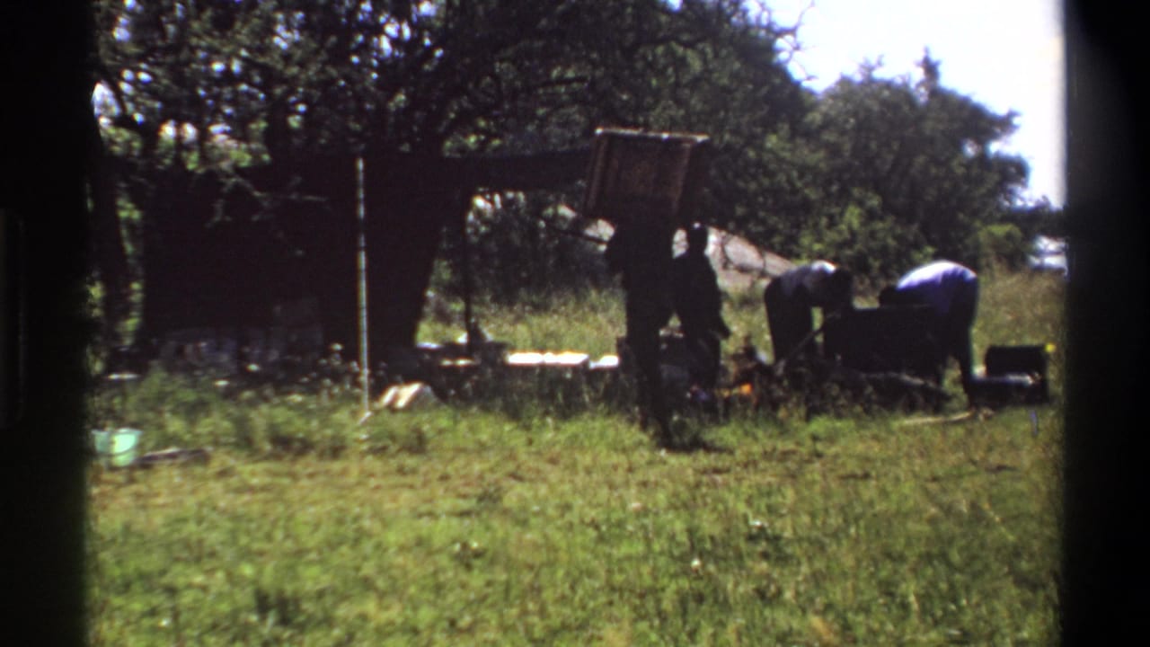 Several men work near a dark chest or load under trees in a grassy area.