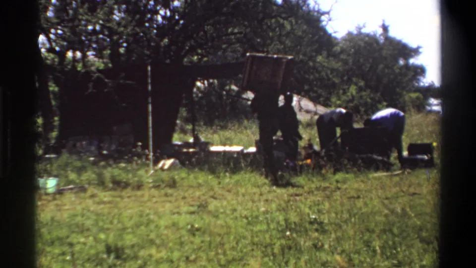 1969 Lagos home-movie frame with men gathered around a heavy dark chest or load under trees in a grassy field.