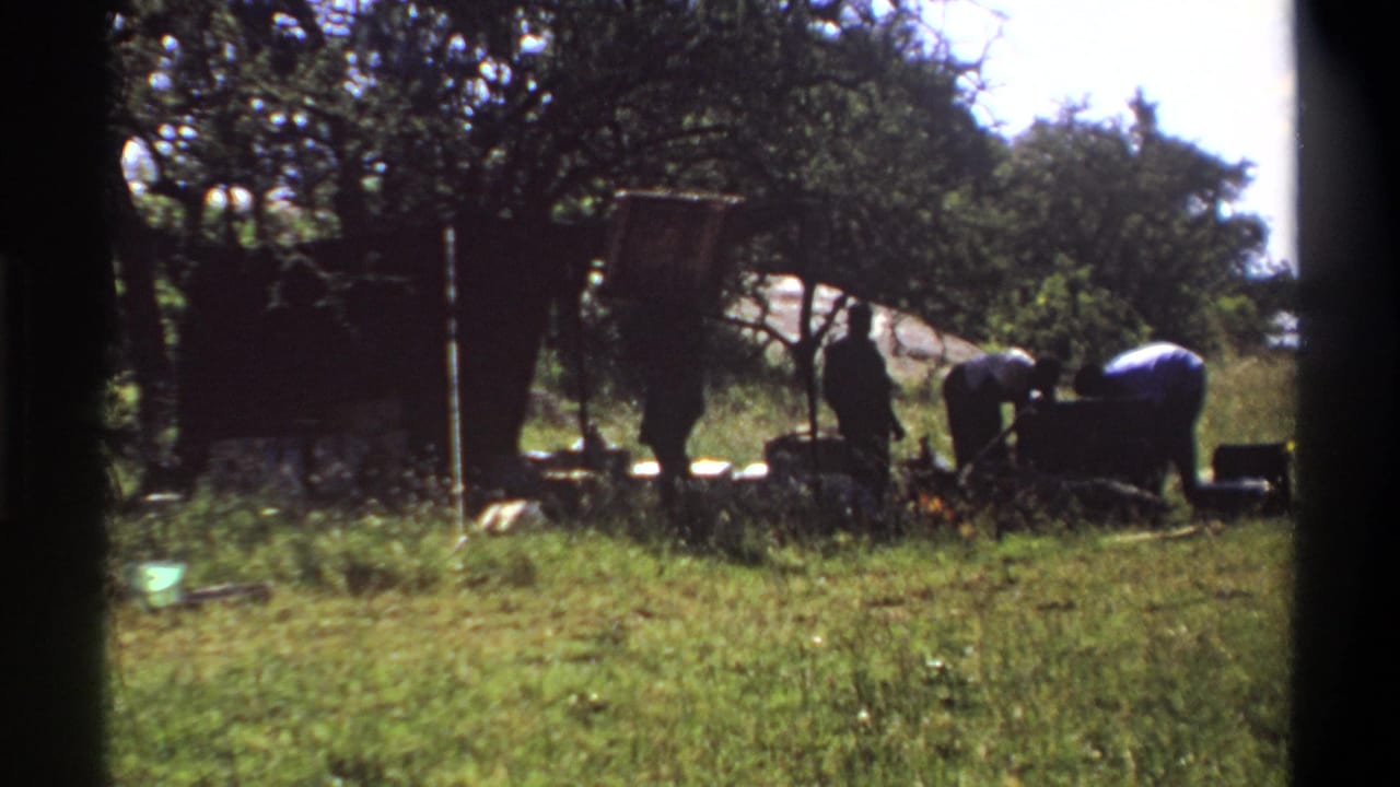 1969 Lagos 8mm frame showing men bent over or standing near a heavy dark load in a sunlit grassy setting.
