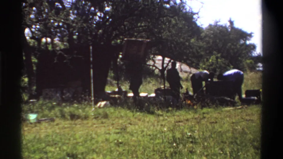 1969 Lagos 8mm frame showing men bent over or standing near a heavy dark load in a sunlit grassy setting.