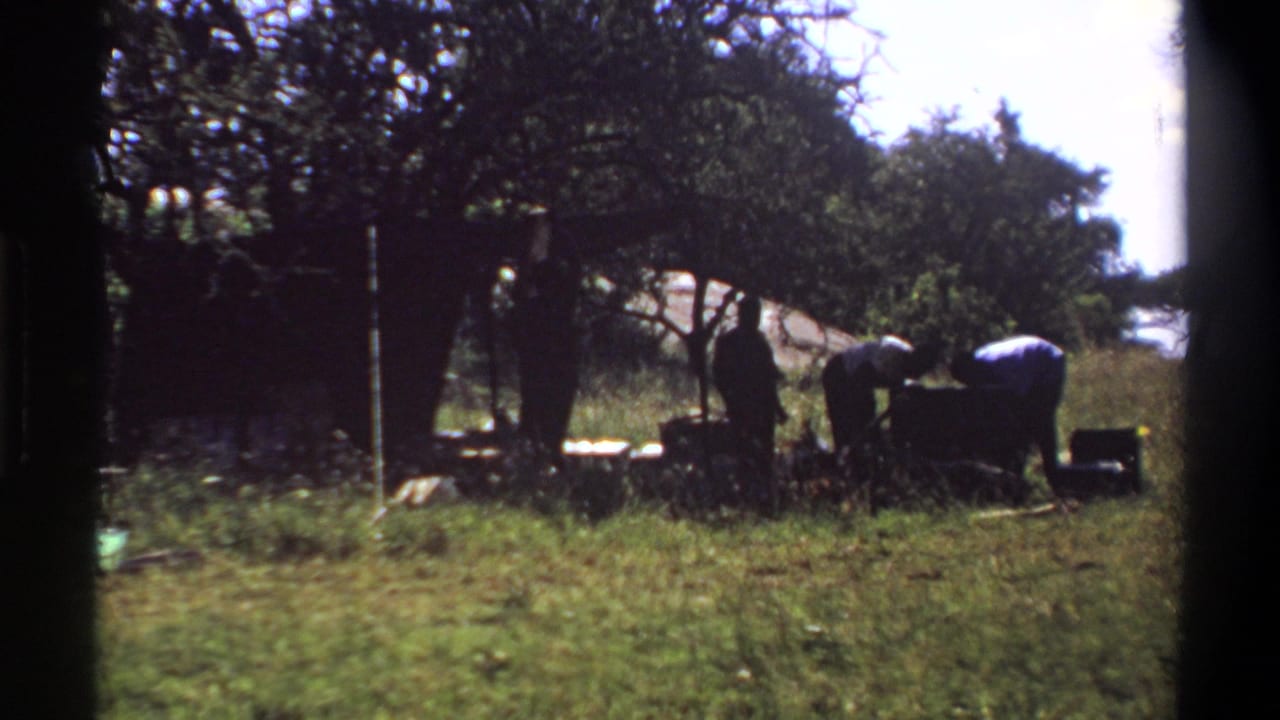 1969 Lagos home-movie frame with men bending around a heavy chest or load in a grassy outdoor area.