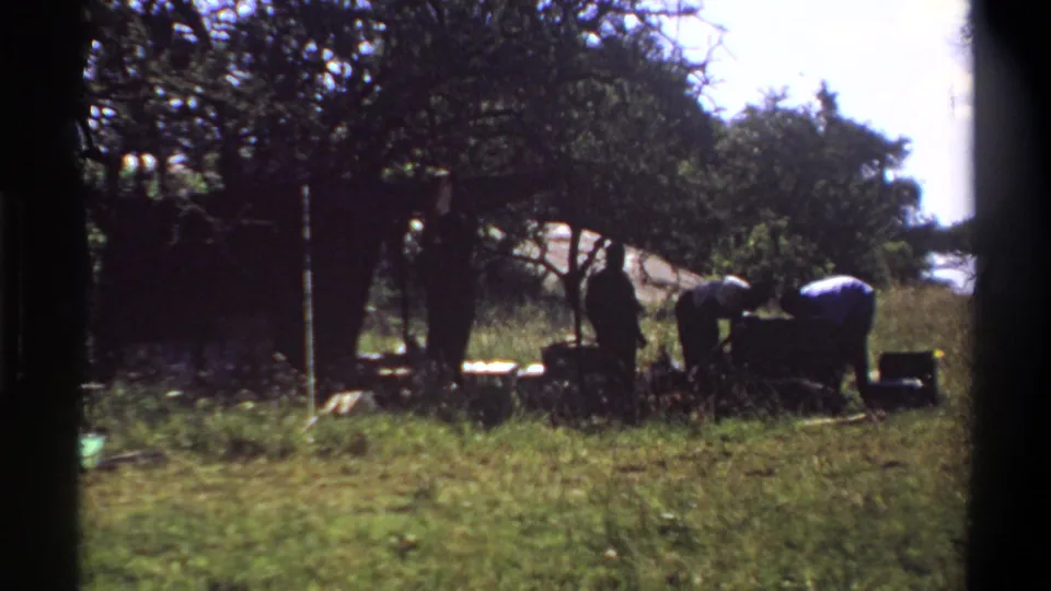 1969 Lagos home-movie frame with men bending around a heavy chest or load in a grassy outdoor area.