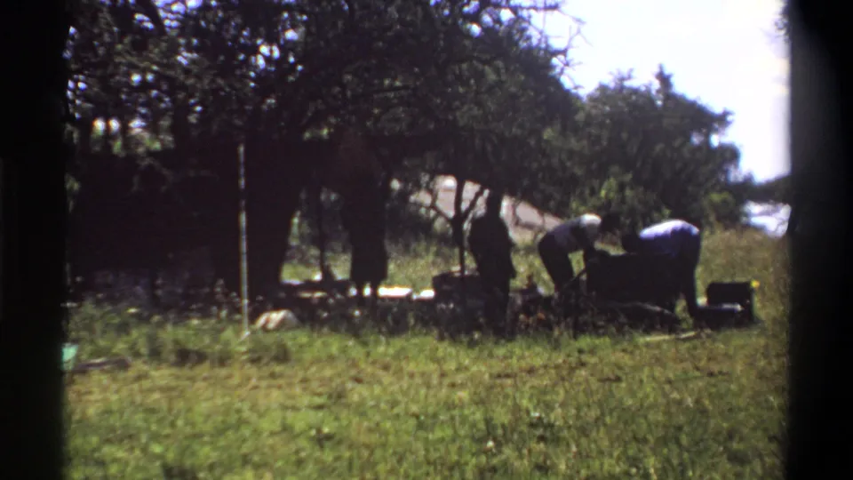1969 Lagos home-movie frame showing people in a grassy shaded campsite area, with two figures bending near a cooking or camp setup.