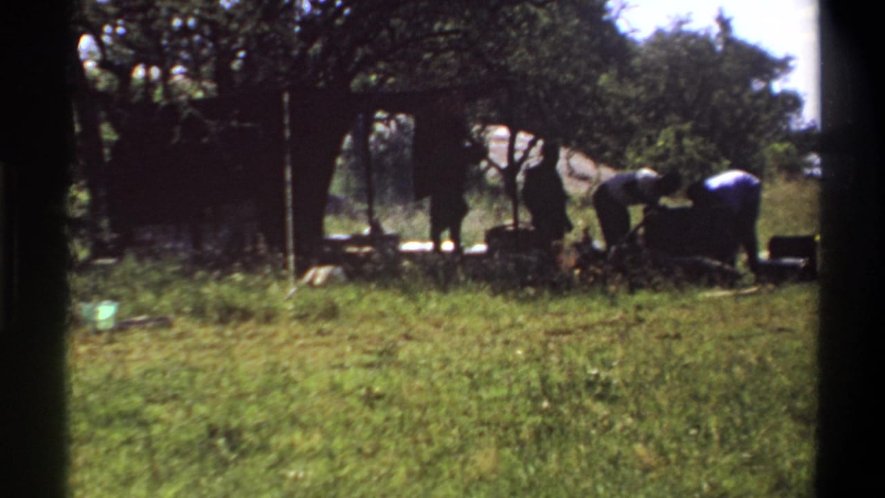 1969 Lagos frame with two people bent over an outdoor camp or cooking area, partly silhouetted under trees.