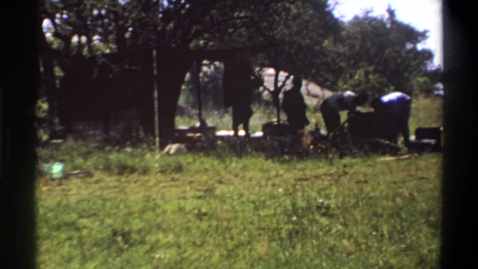 1969 Lagos frame with two people bent over an outdoor camp or cooking area, partly silhouetted under trees.
