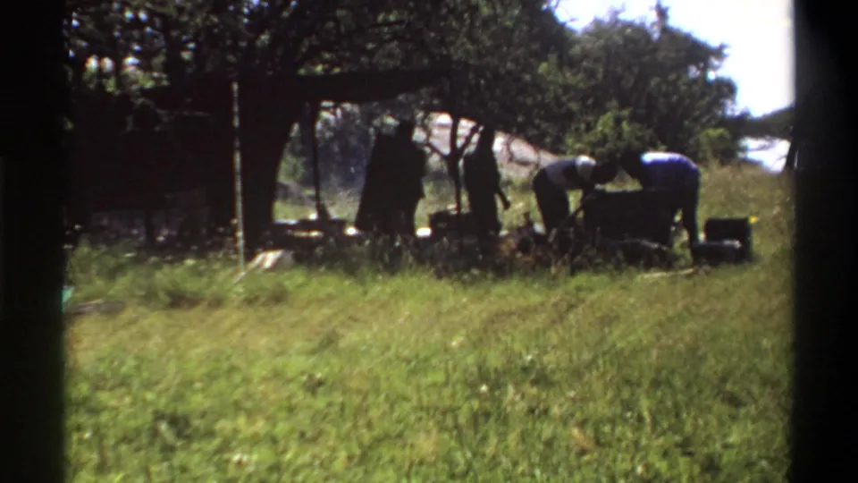 1969 Lagos home-movie still showing a small group around a grassy campsite, with a shelter and trees behind them.