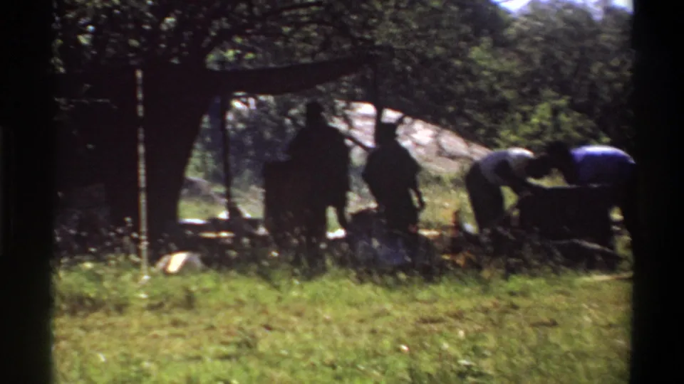 1969 Lagos frame showing silhouetted people under a simple outdoor shelter near grass, trees, and camp equipment.