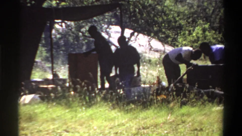 1969 Lagos home-movie frame showing silhouetted figures under a makeshift canopy with pots, grass, trees, and camp activity.