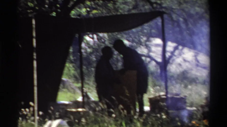 Home-movie frame from Lagos in 1969 showing two silhouetted people near camp gear, smoke, trees, and a shaded canopy.